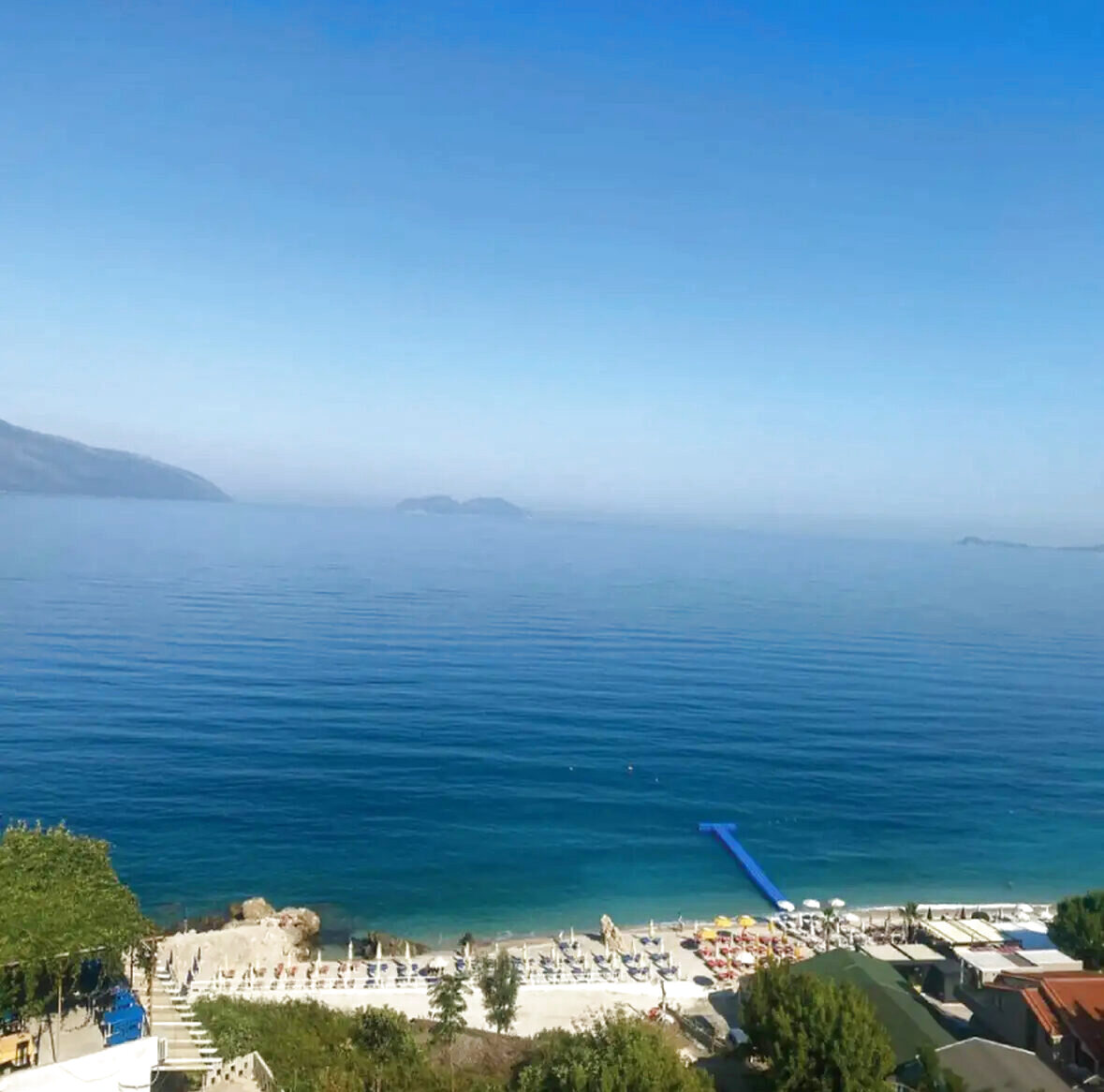 Aerial view of a calm blue bay with a blue floating platform and rows of sunbeds along the shore
