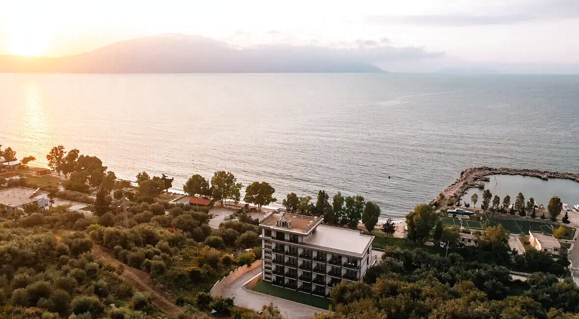 Aerial view of a white modern seafront hotel beside olive groves at sunset, with the sea and a marina beyond