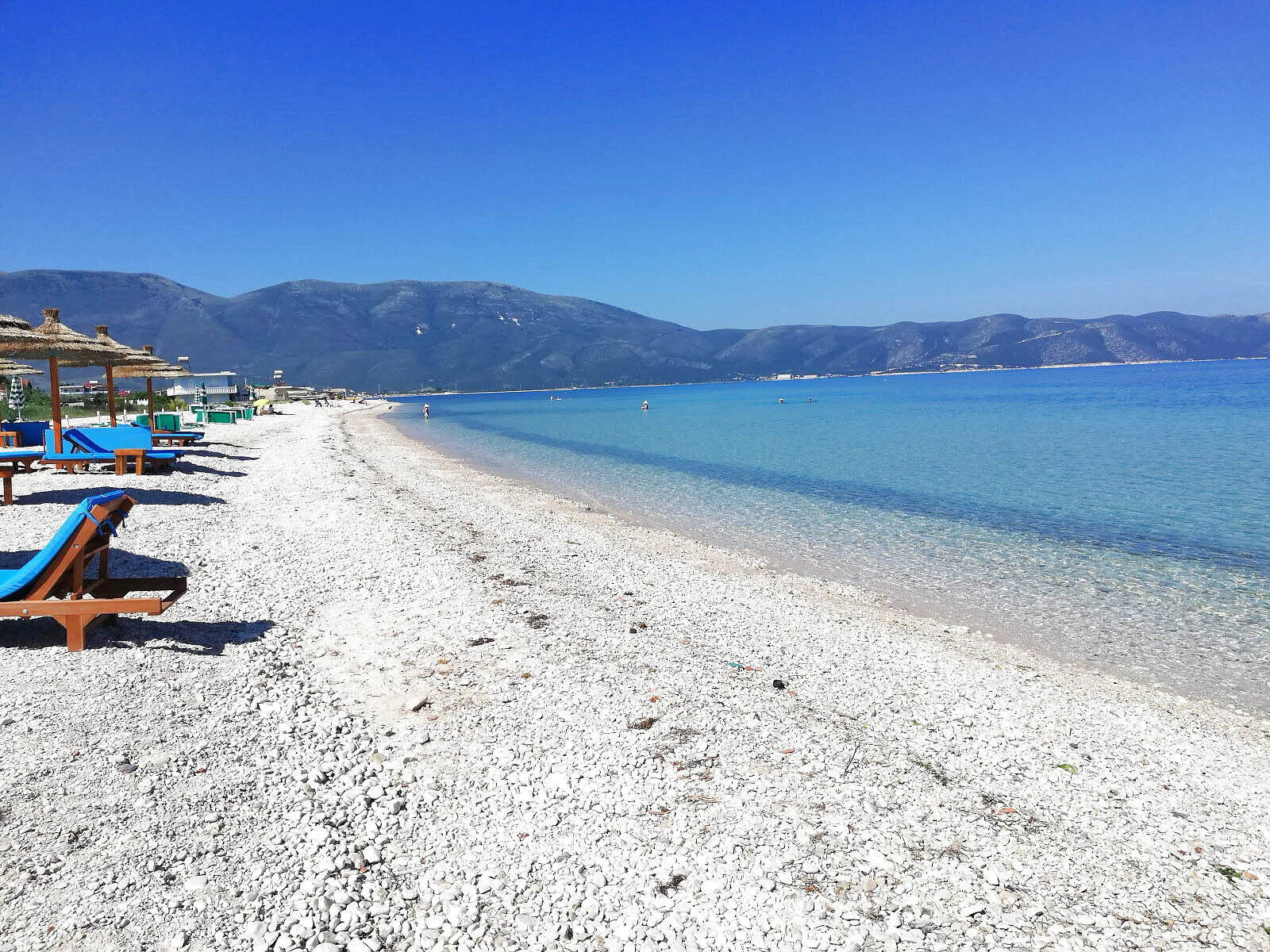 Pebble beach with blue-cushioned wooden loungers and reed parasols beside clear turquoise water
