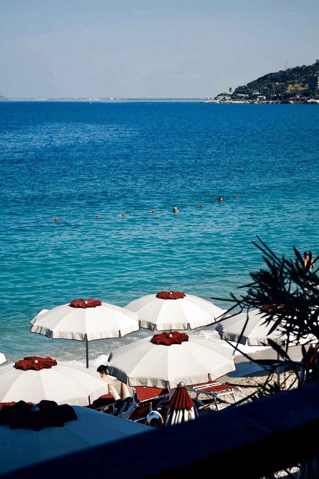 Cream umbrellas with rust-red tops on a shore with a swimmer and a headland