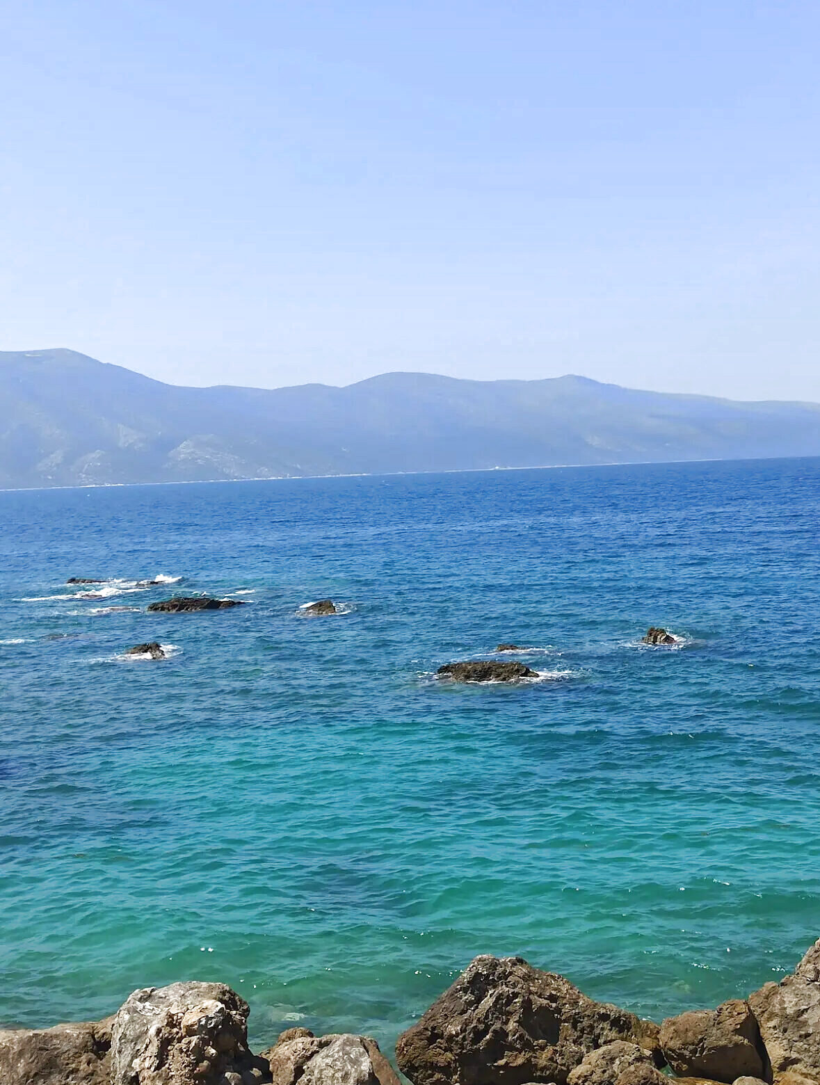 Rocks breaking the surface of a deep blue sea with mountains on the far shore