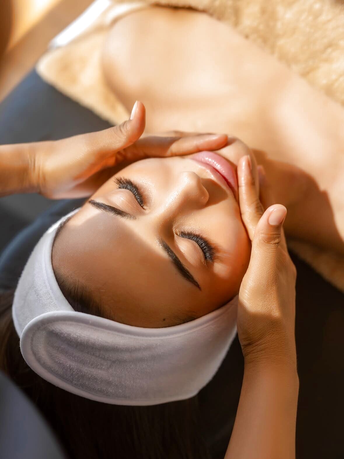 A woman with eyes closed receiving a facial massage, wearing a white headband