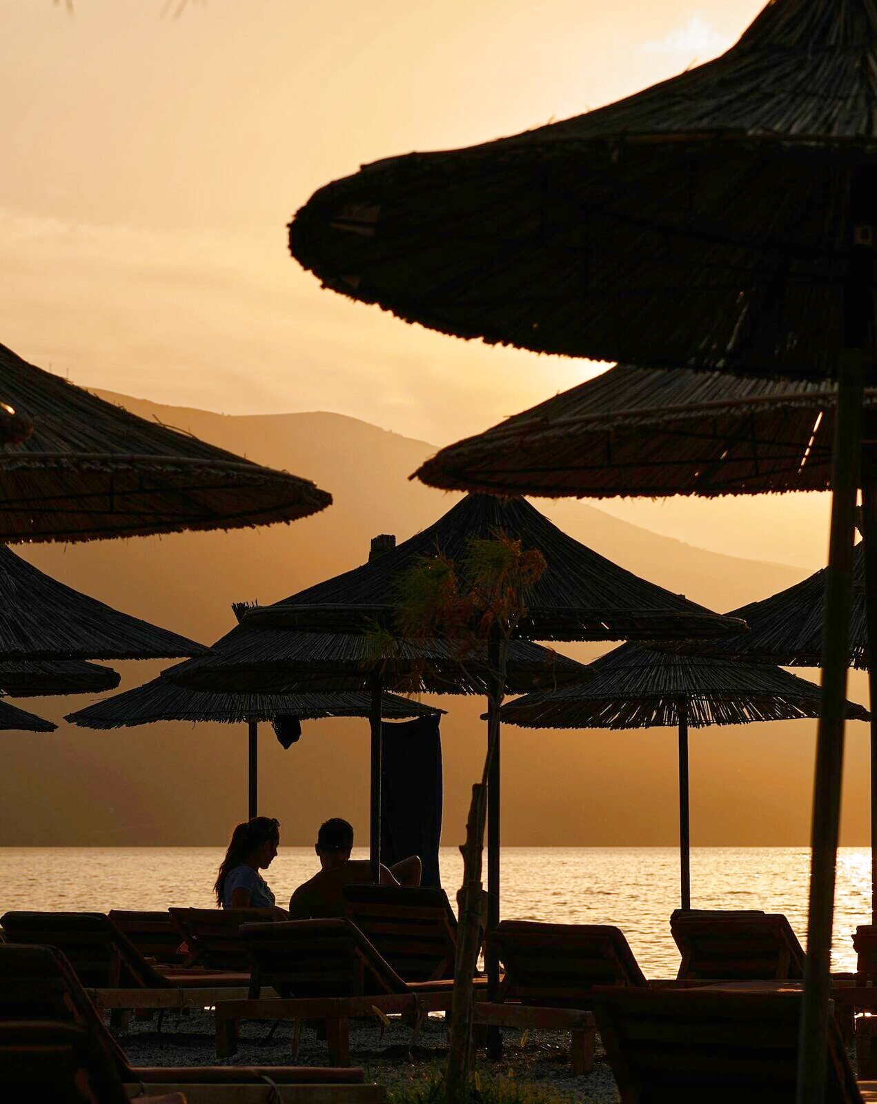 Silhouette of two people talking under reed parasols at sunset with the sea behind