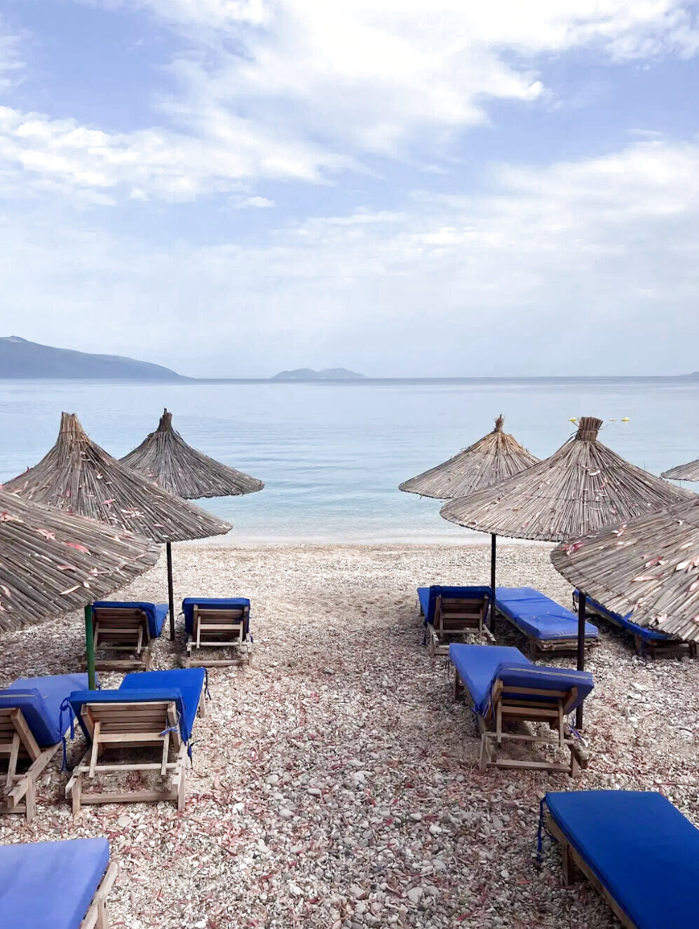 Reed parasols and sunbeds on a sandy shore facing a green mountain ridge across the bay