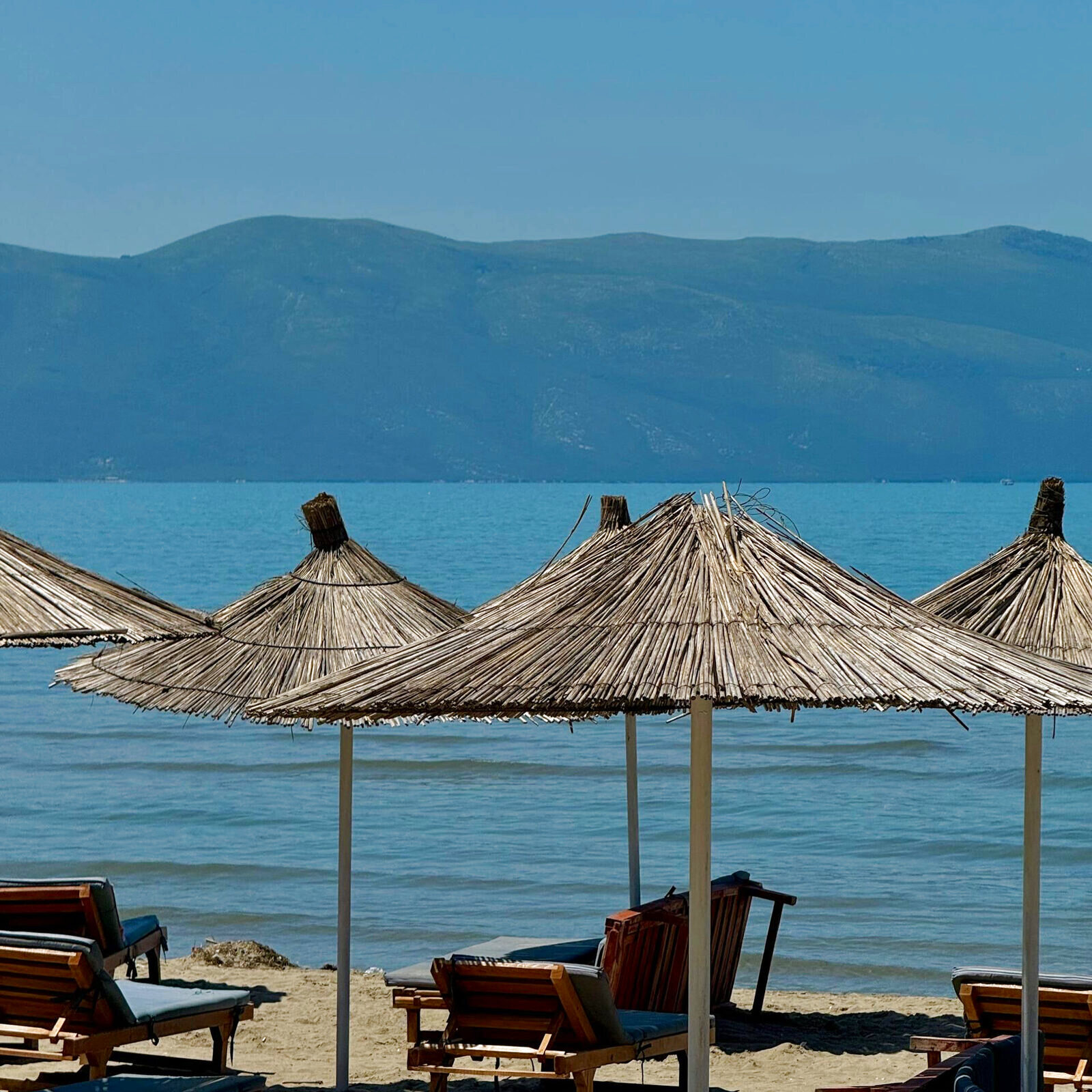 Reed parasols and sunbeds beside a calm sea with mountains on the far shore